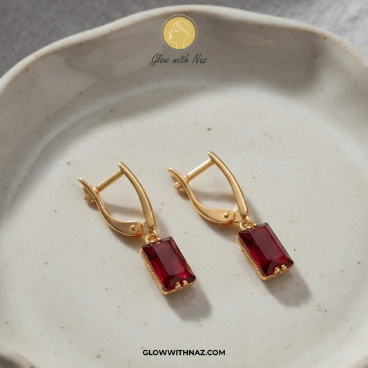 A pair of gold leverback earrings with rectangular deep red gemstones, displayed on a speckled beige ceramic dish. The brand logo 'Glow with Naz' is visible at the top and the website 'GLOWWITHNAZ. studs earrings, small earrings, girls earrings, simple earrings, earrings for girls, black earrings, earrings, ear rings, earrings tops, stud earrings, tops earrings, aesthetic earrings, hoop earrings, drop earrings, diamond earrings, pink earrings, green earrings, ear ring, earrings for women, flower earrings, g