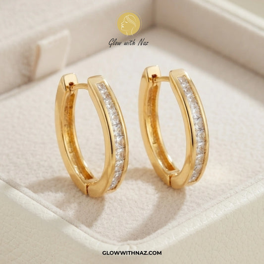 A pair of gold huggie hoop earrings with a channel-set row of clear stones on the front, displayed inside a cream-colored jewelry box. The brand logo 'Glow with Naz' is visible at the top, and the website 'GLOWWITHNAZ. studs earrings, small earrings, girls earrings, simple earrings, earrings for girls, black earrings, earrings, ear rings, earrings tops, stud earrings, tops earrings, aesthetic earrings, hoop earrings, drop earrings, diamond earrings, pink earrings, green earrings, ear ring, earrings for wome