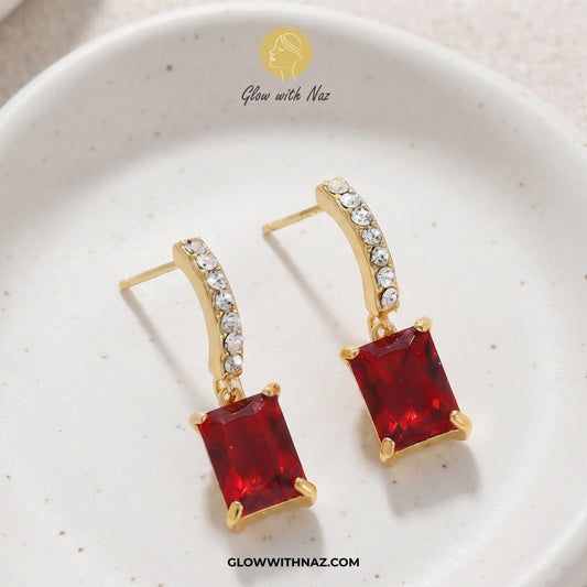 A pair of gold drop earrings with rectangular red gemstones and a curved top set with small clear stones, displayed on a white speckled ceramic dish. The brand name 'Glow with Naz' is visible at the top of the image. studs earrings, small earrings, girls earrings, simple earrings, earrings for girls, black earrings, earrings, ear rings, earrings tops, stud earrings, tops earrings, aesthetic earrings, hoop earrings, drop earrings, diamond earrings, pink earrings, green earrings, ear ring, earrings for women,