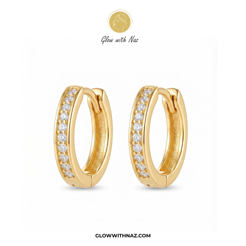 A pair of gold huggie hoop earrings with a row of clear pavé stones on the front, displayed on a clean white background with the brand logo 'Glow with Naz' at the top and the website 'GLOWWITHNAZ.COM' at the bottom. studs earrings, small earrings, girls earrings, simple earrings, earrings for girls, black earrings, earrings, ear rings, earrings tops, stud earrings, tops earrings, aesthetic earrings, hoop earrings, drop earrings, diamond earrings, pink earrings, green earrings, ear ring, earrings for women,