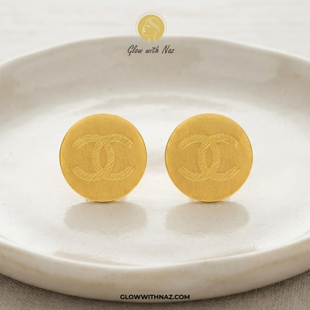 A pair of round gold-tone stud earrings with an embossed interlocking CC logo, displayed on a white ceramic dish. The brand name 'Glow with Naz' is visible at the top, and 'GLOWWITHNAZ. studs earrings, small earrings, girls earrings, simple earrings, earrings for girls, black earrings, earrings, ear rings, earrings tops, stud earrings, tops earrings, aesthetic earrings, hoop earrings, drop earrings, diamond earrings, pink earrings, green earrings, ear ring, earrings for women, flower earrings, golden earrin