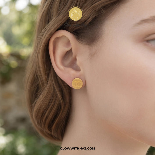 A close-up of a woman's ear wearing a small round gold stud earring, with a matching gold hair clip visible in her hair. The background is softly blurred outdoor greenery. studs earrings, small earrings, girls earrings, simple earrings, earrings for girls, black earrings, earrings, ear rings, earrings tops, stud earrings, tops earrings, aesthetic earrings, hoop earrings, drop earrings, diamond earrings, pink earrings, green earrings, ear ring, earrings for women, flower earrings, golden earrings, bali earri