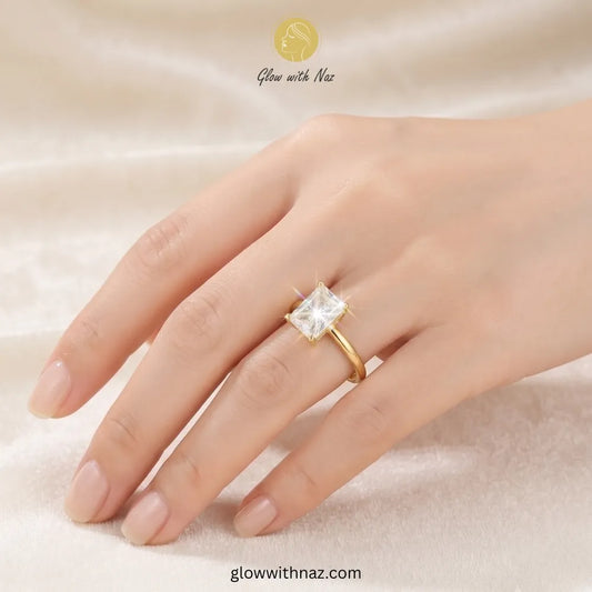 A gold ring with a square clear gemstone set on a person's ring finger, shown against a soft beige fabric background. The brand name 'Glow with Naz' is visible at the top. ring, girls rings, rings for girls, rings simple, simple rings, stainless steel jewelry, jewellery set, aesthetic jewelry, simple jewellery, chain, jewelry, artificial jewellery
