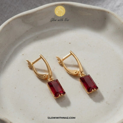 A pair of gold leverback earrings with rectangular deep red gemstones, displayed on a speckled beige ceramic dish. The brand logo 'Glow with Naz' is visible at the top and the website 'GLOWWITHNAZ. studs earrings, small earrings, girls earrings, simple earrings, earrings for girls, black earrings, earrings, ear rings, earrings tops, stud earrings, tops earrings, aesthetic earrings, hoop earrings, drop earrings, diamond earrings, pink earrings, green earrings, ear ring, earrings for women, flower earrings, g