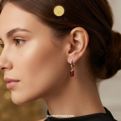 A close-up side profile of a woman wearing a small gold hoop earring with a dangling rectangular red gemstone. The image features a gold coin in her hair for scale and a watermark 'GLOWWITHNAZ. studs earrings, small earrings, girls earrings, simple earrings, earrings for girls, black earrings, earrings, ear rings, earrings tops, stud earrings, tops earrings, aesthetic earrings, hoop earrings, drop earrings, diamond earrings, pink earrings, green earrings, ear ring, earrings for women, flower earrings, golde