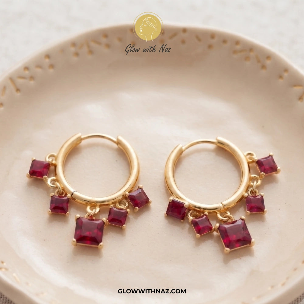 A pair of gold huggie hoop earrings with dangling red gemstone charms, displayed on a beige ceramic dish. The brand logo 'Glow with Naz' is visible at the top and the website URL at the bottom. studs earrings, small earrings, girls earrings, simple earrings, earrings for girls, black earrings, earrings, ear rings, earrings tops, stud earrings, tops earrings, aesthetic earrings, hoop earrings, drop earrings, diamond earrings, pink earrings, green earrings, ear ring, earrings for women, flower earrings, golde