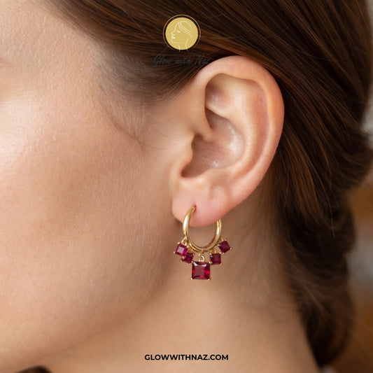 A close-up of a model's ear wearing a small gold hoop earring adorned with several square-cut dark red gemstones. A coin is shown near the top for scale. studs earrings, small earrings, girls earrings, simple earrings, earrings for girls, black earrings, earrings, ear rings, earrings tops, stud earrings, tops earrings, aesthetic earrings, hoop earrings, drop earrings, diamond earrings, pink earrings, green earrings, ear ring, earrings for women, flower earrings, golden earrings, bali earrings, ear jewellery