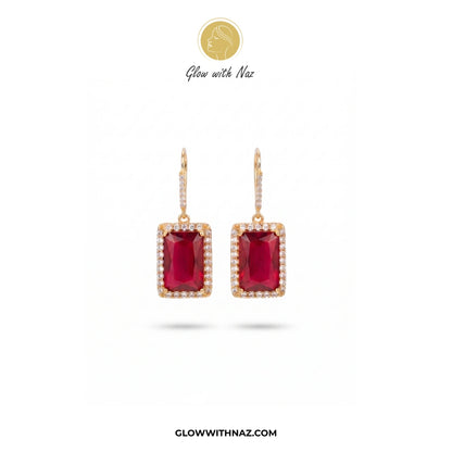 A pair of gold drop earrings featuring rectangular emerald-cut deep red ruby centers, surrounded by a halo of small clear pavé stones, shown on a white background with the brand logo 'Glow with Naz' at the top. studs earrings, small earrings, girls earrings, simple earrings, earrings for girls, black earrings, earrings, ear rings, earrings tops, stud earrings, tops earrings, aesthetic earrings, hoop earrings, drop earrings, diamond earrings, pink earrings, green earrings, ear ring, earrings for women, flowe