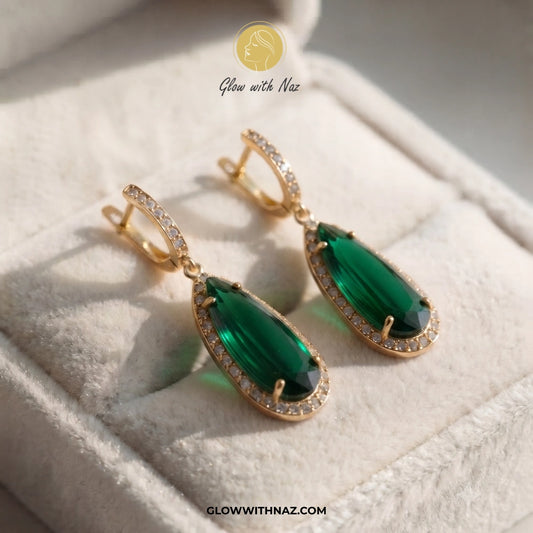 A pair of gold drop earrings with large emerald-cut green gemstones and small clear stones along the border, displayed on a soft cream jewelry box. A watermark logo for Glow with Naz is visible at the top, and the website CLOWWITNAZ. studs earrings, small earrings, girls earrings, simple earrings, earrings for girls, black earrings, earrings, ear rings, earrings tops, stud earrings, tops earrings, aesthetic earrings, hoop earrings, drop earrings, diamond earrings, pink earrings, green earrings, ear ring, ea
