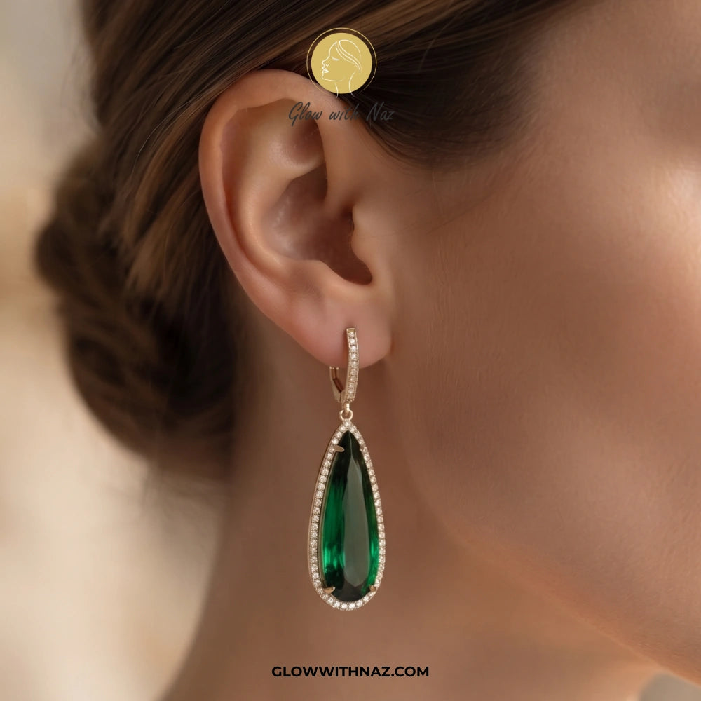 A close-up of a model's ear wearing a teardrop-shaped green gemstone drop earring with a silver-toned border and small clear accent stones. A gold coin is placed near the ear for scale. studs earrings, small earrings, girls earrings, simple earrings, earrings for girls, black earrings, earrings, ear rings, earrings tops, stud earrings, tops earrings, aesthetic earrings, hoop earrings, drop earrings, diamond earrings, pink earrings, green earrings, ear ring, earrings for women, flower earrings, golden earrin