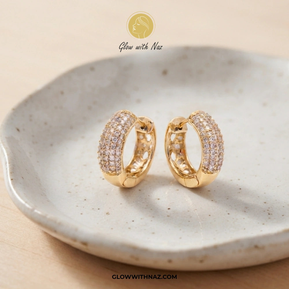 A pair of gold huggie hoop earrings paved with small clear stones, displayed on a speckled ceramic dish. The brand logo 'Glow with Naz' is visible at the top and the website 'GLOWWITHNAZ.