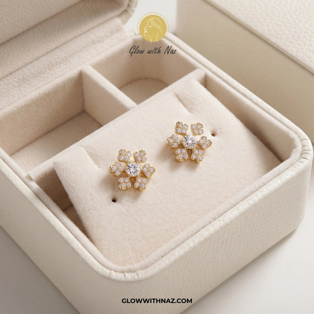 A pair of gold flower-shaped stud earrings with clear gemstones, displayed on a cream-colored jewelry box cushion. The brand logo 'Glow with Naz' is visible at the top. studs earrings, small earrings, girls earrings, simple earrings, earrings for girls, black earrings, earrings, ear rings, earrings tops, stud earrings, tops earrings, aesthetic earrings, hoop earrings, drop earrings, diamond earrings, pink earrings, green earrings, ear ring, earrings for women, flower earrings, golden earrings, bali earrings