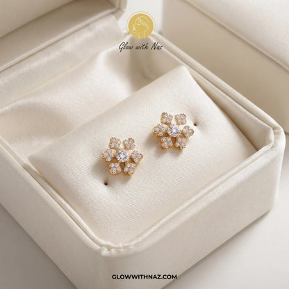 A pair of gold flower-shaped stud earrings with clear stones, displayed on a cream satin-lined jewelry box. A watermark logo for 'Glow with Naz' is visible at the top, and the URL 'CLOWWITHNAZ. studs earrings, small earrings, girls earrings, simple earrings, earrings for girls, black earrings, earrings, ear rings, earrings tops, stud earrings, tops earrings, aesthetic earrings, hoop earrings, drop earrings, diamond earrings, pink earrings, green earrings, ear ring, earrings for women, flower earrings, golde