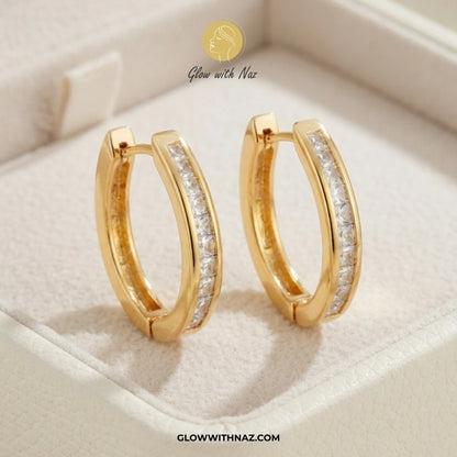 A pair of gold huggie hoop earrings with a channel-set row of clear stones on the front, displayed inside a cream-colored jewelry box. The brand logo 'Glow with Naz' is visible at the top, and the website 'GLOWWITHNAZ. studs earrings, small earrings, girls earrings, simple earrings, earrings for girls, black earrings, earrings, ear rings, earrings tops, stud earrings, tops earrings, aesthetic earrings, hoop earrings, drop earrings, diamond earrings, pink earrings, green earrings, ear ring, earrings for wome