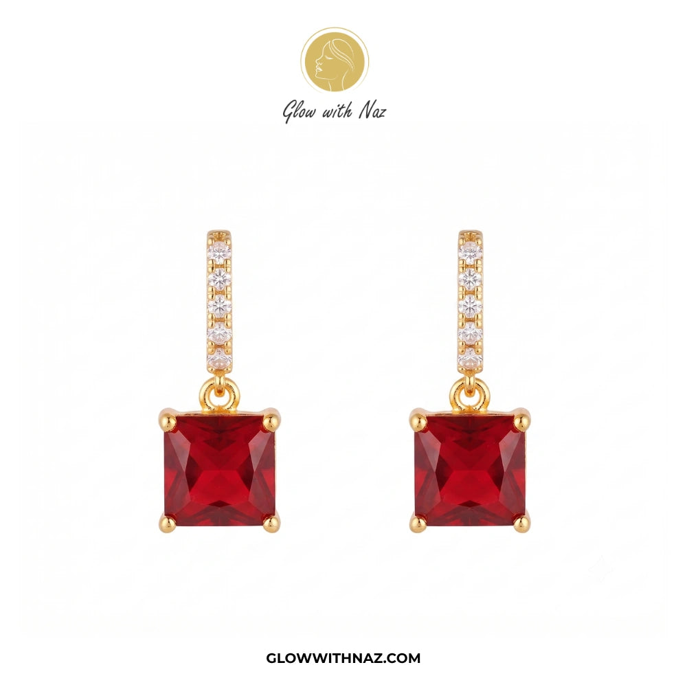 A pair of gold drop earrings featuring square-cut red gemstones, with a vertical bar of small clear stones above each red stone. The brand logo 'Glow with Naz' is visible at the top and the website 'GLOWWITHNAZ. studs earrings, small earrings, girls earrings, simple earrings, earrings for girls, black earrings, earrings, ear rings, earrings tops, stud earrings, tops earrings, aesthetic earrings, hoop earrings, drop earrings, diamond earrings, pink earrings, green earrings, ear ring, earrings for women, flow