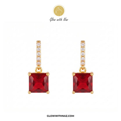 A pair of gold drop earrings featuring square-cut red gemstones, with a vertical bar of small clear stones above each red stone. The brand logo 'Glow with Naz' is visible at the top and the website 'GLOWWITHNAZ. studs earrings, small earrings, girls earrings, simple earrings, earrings for girls, black earrings, earrings, ear rings, earrings tops, stud earrings, tops earrings, aesthetic earrings, hoop earrings, drop earrings, diamond earrings, pink earrings, green earrings, ear ring, earrings for women, flow