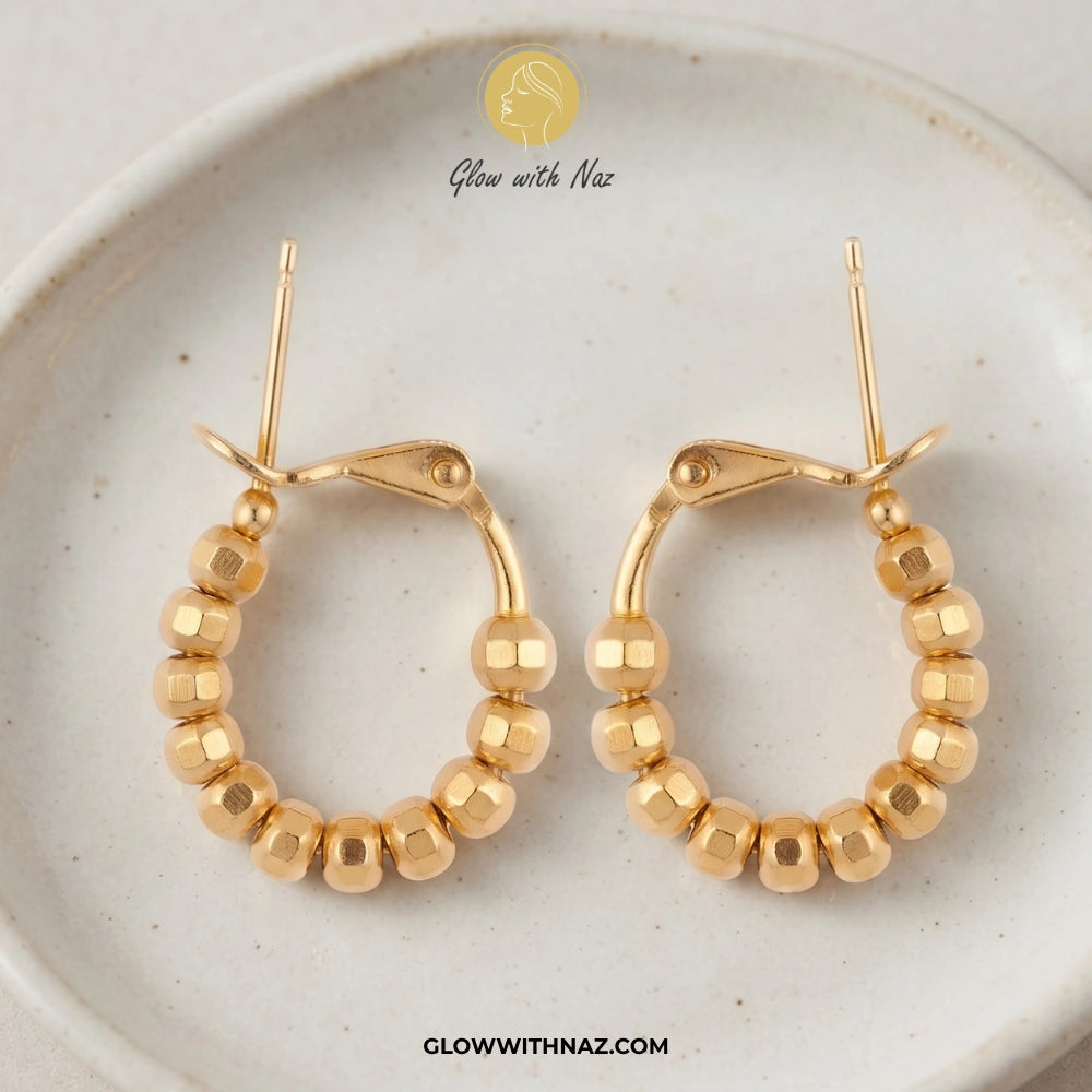 A pair of gold hoop earrings with faceted bead details, displayed on a light beige ceramic plate. A brand logo for 'Glow with Naz' is visible at the top, and a website URL is visible at the bottom. studs earrings, small earrings, girls earrings, simple earrings, earrings for girls, black earrings, earrings, ear rings, earrings tops, stud earrings, tops earrings, aesthetic earrings, hoop earrings, drop earrings, diamond earrings, pink earrings, green earrings, ear ring, earrings for women, flower earrings, g