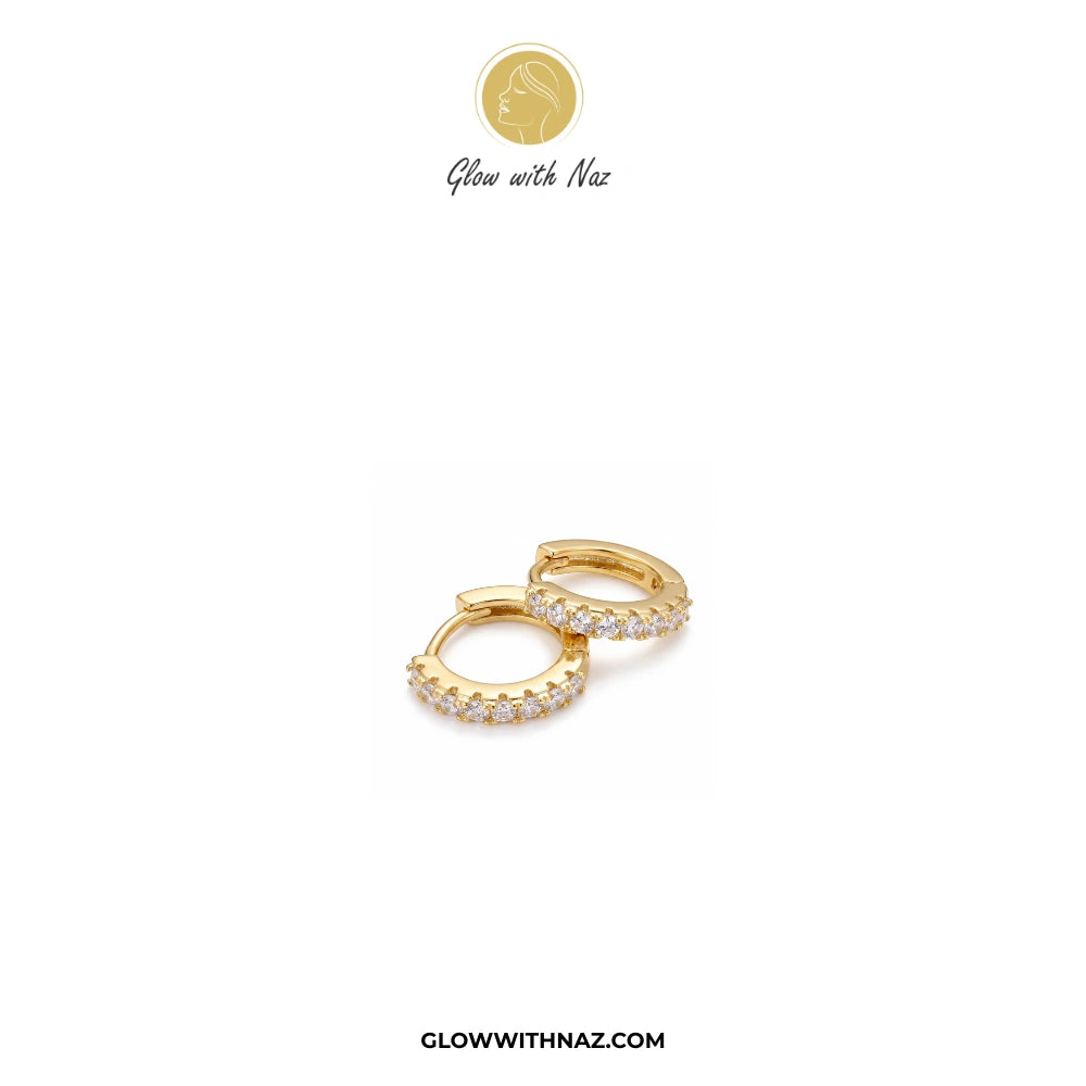 A pair of gold huggie hoop earrings adorned with small clear pavé stones, displayed on a clean white background with the brand logo 'Glow with Naz' at the top and 'GLOWWITHNAZ.COM' at the bottom.studs earrings, small earrings, girls earrings, simple earrings, earrings for girls, black earrings, earrings, ear rings, earrings tops, stud earrings, tops earrings, aesthetic earrings, hoop earrings, drop earrings, diamond earrings, pink earrings, green earrings, ear ring, earrings for women, flower earrings, gold