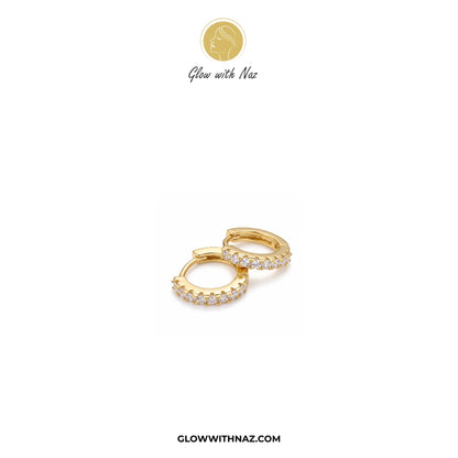 A pair of gold huggie hoop earrings adorned with small clear pavé stones, displayed on a clean white background with the brand logo 'Glow with Naz' at the top and 'GLOWWITHNAZ.COM' at the bottom.studs earrings, small earrings, girls earrings, simple earrings, earrings for girls, black earrings, earrings, ear rings, earrings tops, stud earrings, tops earrings, aesthetic earrings, hoop earrings, drop earrings, diamond earrings, pink earrings, green earrings, ear ring, earrings for women, flower earrings, gold