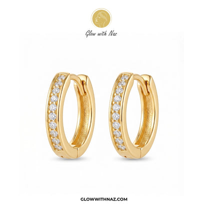 A pair of gold huggie hoop earrings with a row of clear pavé stones on the front, displayed on a clean white background with the brand logo 'Glow with Naz' at the top and the website 'GLOWWITHNAZ.COM' at the bottom. studs earrings, small earrings, girls earrings, simple earrings, earrings for girls, black earrings, earrings, ear rings, earrings tops, stud earrings, tops earrings, aesthetic earrings, hoop earrings, drop earrings, diamond earrings, pink earrings, green earrings, ear ring, earrings for women, 