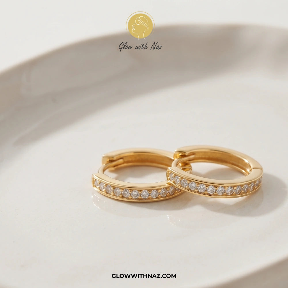 A pair of gold huggie hoop earrings with a row of small clear pavé stones, resting on a white ceramic dish. The brand logo 'Glow with Naz' is visible at the top, and the website 'GLOWWITHNAZ. studs earrings, small earrings, girls earrings, simple earrings, earrings for girls, black earrings, earrings, ear rings, earrings tops, stud earrings, tops earrings, aesthetic earrings, hoop earrings, drop earrings, diamond earrings, pink earrings, green earrings, ear ring, earrings for women, flower earrings, golden 