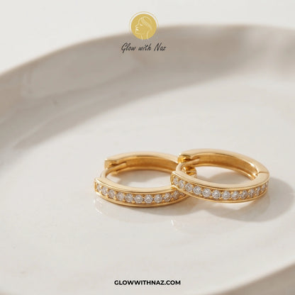 A pair of gold huggie hoop earrings with a row of small clear pavé stones, resting on a white ceramic dish. The brand logo 'Glow with Naz' is visible at the top, and the website 'GLOWWITHNAZ. studs earrings, small earrings, girls earrings, simple earrings, earrings for girls, black earrings, earrings, ear rings, earrings tops, stud earrings, tops earrings, aesthetic earrings, hoop earrings, drop earrings, diamond earrings, pink earrings, green earrings, ear ring, earrings for women, flower earrings, golden 
