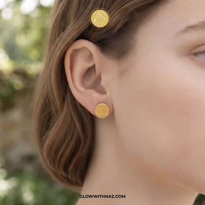A close-up of a woman's ear wearing a small round gold stud earring, with a matching gold hair clip visible in her hair. The background is softly blurred outdoor greenery. studs earrings, small earrings, girls earrings, simple earrings, earrings for girls, black earrings, earrings, ear rings, earrings tops, stud earrings, tops earrings, aesthetic earrings, hoop earrings, drop earrings, diamond earrings, pink earrings, green earrings, ear ring, earrings for women, flower earrings, golden earrings, bali earri
