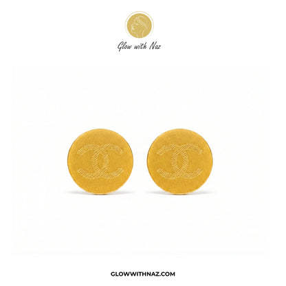 A pair of round gold-tone Chanel stud earrings with the interlocking CC logo embossed on the front, displayed on a clean white background with Glow with Naz branding. studs earrings, small earrings, girls earrings, simple earrings, earrings for girls, black earrings, earrings, ear rings, earrings tops, stud earrings, tops earrings, aesthetic earrings, hoop earrings, drop earrings, diamond earrings, pink earrings, green earrings, ear ring, earrings for women, flower earrings, golden earrings, bali earrings, 