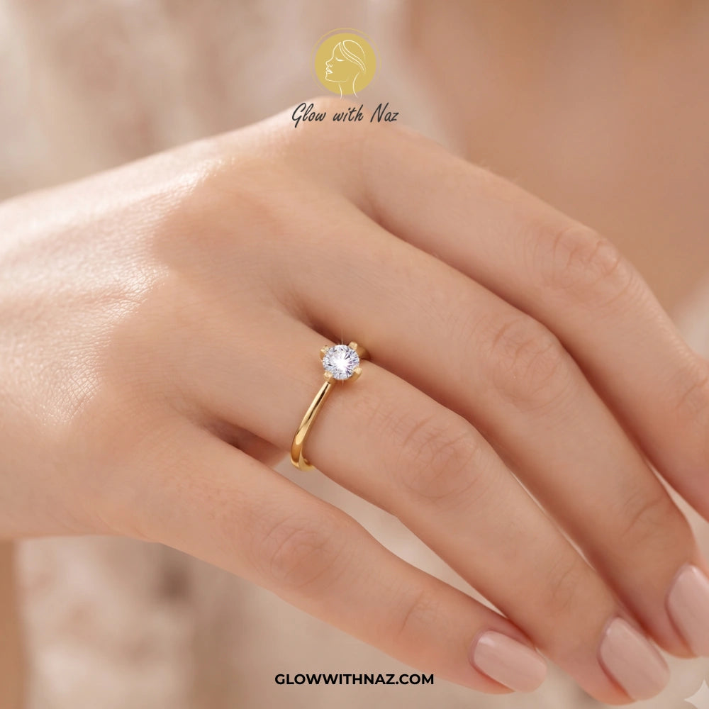 A gold solitaire ring with a clear round gemstone worn on a woman's finger. The image features a warm, soft-focus background and a brand watermark 'Glow with Naz' at the top and 'GLOWWITHNAZ. ring, girls rings, rings for girls, rings simple, simple rings, stainless steel jewelry, jewellery set, aesthetic jewelry, simple jewellery, chain, jewelry, artificial jewellery