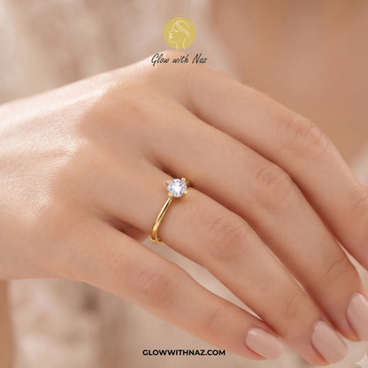 A gold solitaire ring with a clear round gemstone worn on a woman's finger. The image features a warm, soft-focus background and a brand watermark 'Glow with Naz' at the top and 'GLOWWITHNAZ. ring, girls rings, rings for girls, rings simple, simple rings, stainless steel jewelry, jewellery set, aesthetic jewelry, simple jewellery, chain, jewelry, artificial jewellery