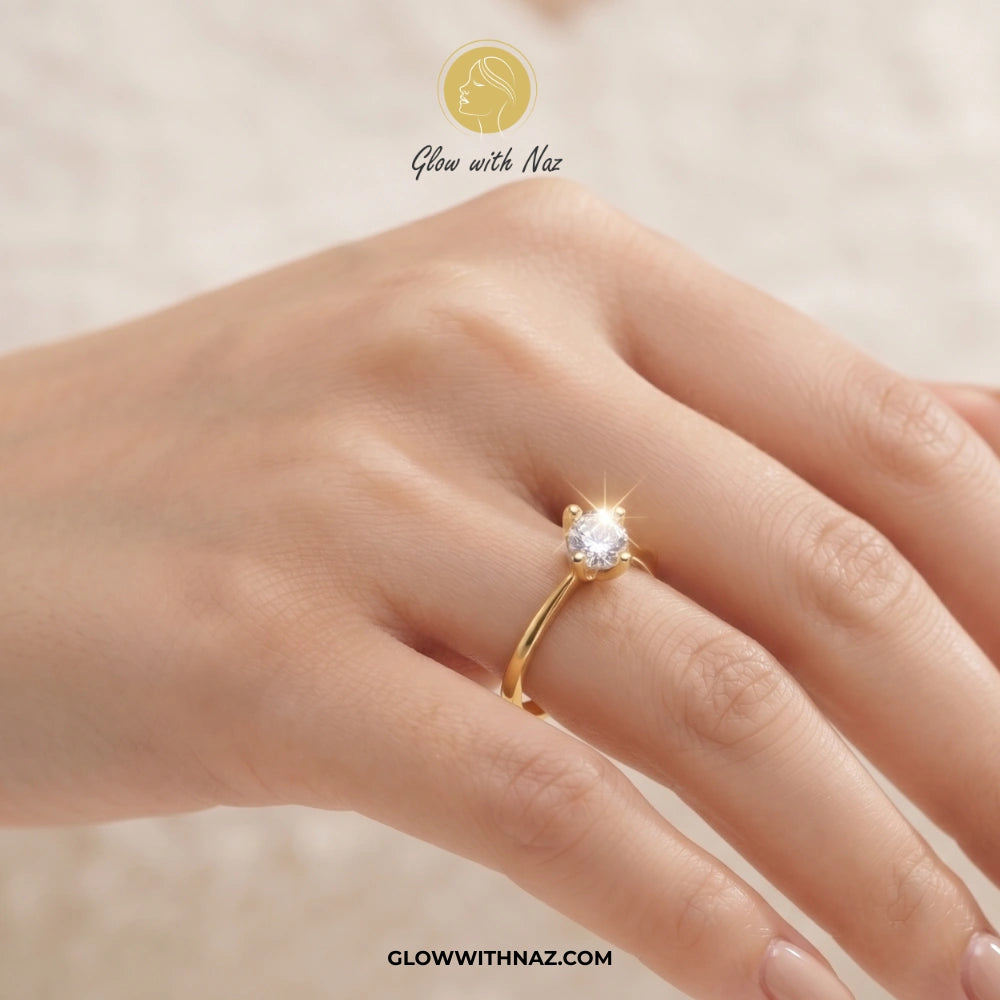 A close-up of a hand wearing a gold solitaire ring with a clear gemstone. The image features a 'Glow with Naz' logo at the top and the website 'GLOWWITHNAZ. ring, girls rings, rings for girls, rings simple, simple rings, stainless steel jewelry, jewellery set, aesthetic jewelry, simple jewellery, chain, jewelry, artificial jewellery