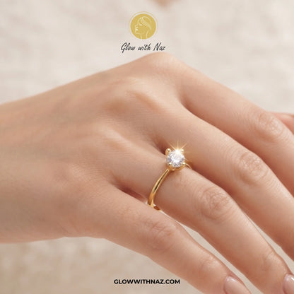 A close-up of a hand wearing a gold solitaire ring with a clear gemstone. The image features a 'Glow with Naz' logo at the top and the website 'GLOWWITHNAZ. ring, girls rings, rings for girls, rings simple, simple rings, stainless steel jewelry, jewellery set, aesthetic jewelry, simple jewellery, chain, jewelry, artificial jewellery