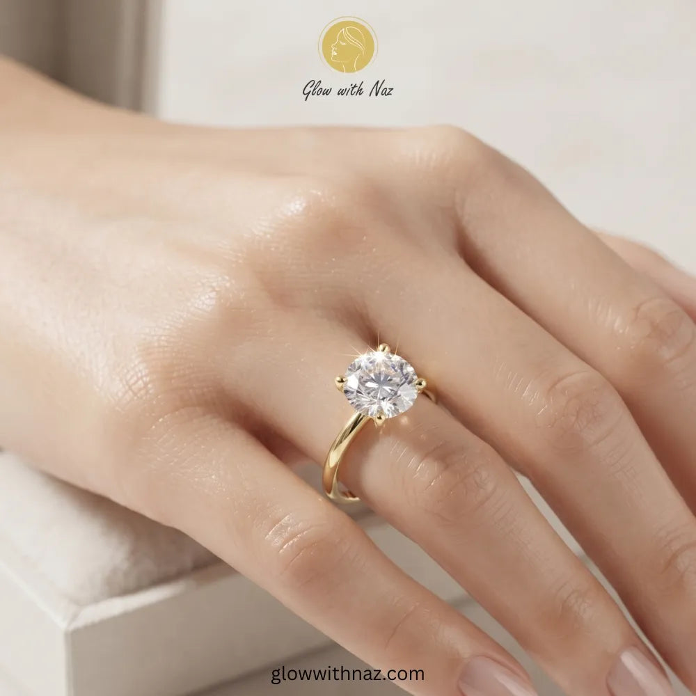 A close-up of a hand wearing a yellow gold solitaire ring with a clear round-cut gemstone. The brand logo 'Glow with Naz' is visible at the top of the image. ring, girls rings, rings for girls, rings simple, simple rings, stainless steel jewelry, jewellery set, aesthetic jewelry, simple jewellery, chain, jewelry, artificial jewellery