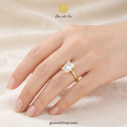 A gold ring with a square clear gemstone set on a person's ring finger, shown against a soft beige fabric background. The brand name 'Glow with Naz' is visible at the top. ring, girls rings, rings for girls, rings simple, simple rings, stainless steel jewelry, jewellery set, aesthetic jewelry, simple jewellery, chain, jewelry, artificial jewellery