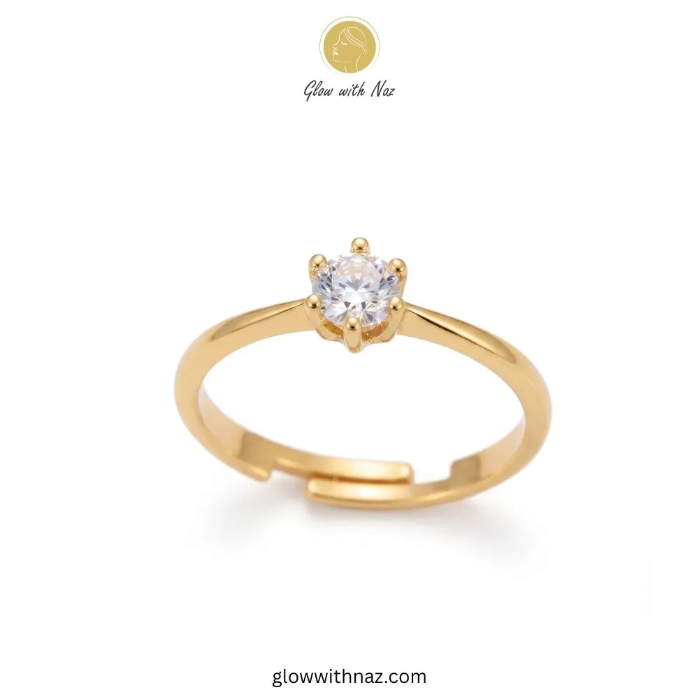 A gold adjustable ring with a single round clear gemstone in a prong setting, shown on a white background with a Glow with Naz logo at the top and glowwithnaz.com at the bottom. ring, girls rings, rings for girls, rings simple, simple rings, stainless steel jewelry, jewellery set, aesthetic jewelry, simple jewellery, chain, jewelry, artificial jewellery