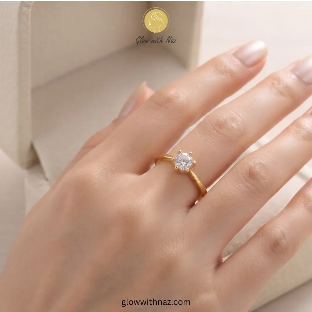 A close-up of a hand wearing a yellow gold ring with a single round-cut clear gemstone. The background shows a soft beige jewelry box, with the brand name 'Glow with Naz'  ring, girls rings, rings for girls, rings simple, simple rings, stainless steel jewelry, jewellery set, aesthetic jewelry, simple jewellery, chain, jewelry, artificial jewelleryvisible.