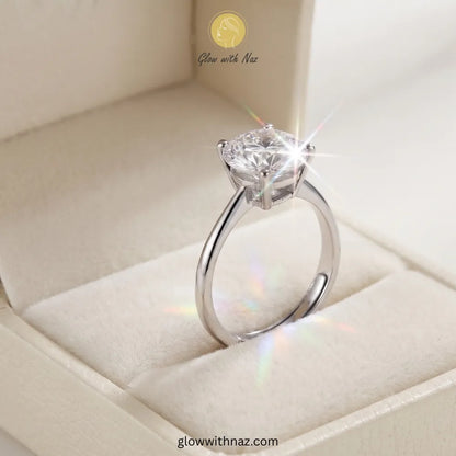 A silver solitaire engagement ring with a round brilliant-cut diamond, displayed inside a cream-colored velvet ring box. The image features a stylized starburst and rainbow light flare effect around the diamond, with the brand name 'Glow with Naz' visible in the upper portion. ring, girls rings, rings for girls, rings simple, simple rings, stainless steel jewelry, jewellery set, aesthetic jewelry, simple jewellery, chain, jewelry, artificial jewellery