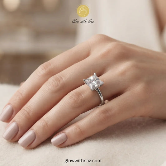 A silver ring with a clear princess-cut gemstone worn on a hand with light pink manicured nails, set against a soft beige background with a 'Glow with Naz' watermark. ring, girls rings, rings for girls, rings simple, simple rings, stainless steel jewelry, jewellery set, aesthetic jewelry, simple jewellery, chain, jewelry, artificial jewellery