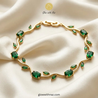 A gold leaf-shaped bracelet adorned with green marquise and oval gemstones, displayed on a draped cream satin fabric. The brand logo 'Glow with Naz' is visible at the top. Tulip Rapunzel bracelet, floral tulip bracelet, Rapunzel inspired jewelry, aesthetic tulip bracelet, trendy charm bracelet, stainless steel tulip bracelet, gold plated flower bracelet, delicate floral wrist accessory, cute aesthetic jewelry, viral tulip bracelet, Rapunzel theme accessories, dainty floral bracelet, crystal tulip charm, sta