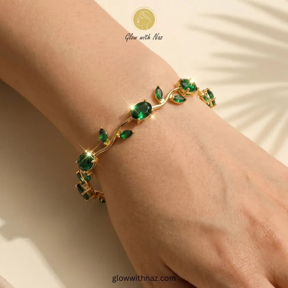 A gold bracelet with green gemstone leaves and oval emerald-cut stones, shown worn on a person's wrist against a warm beige background with soft leaf shadows. Tulip Rapunzel bracelet, floral tulip bracelet, Rapunzel inspired jewelry, aesthetic tulip bracelet, trendy charm bracelet, stainless steel tulip bracelet, gold plated flower bracelet, delicate floral wrist accessory, cute aesthetic jewelry, viral tulip bracelet, Rapunzel theme accessories, dainty floral bracelet, crystal tulip charm, stacking bracele
