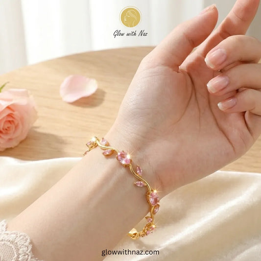 A gold-toned floral vine bracelet with pink rhinestones worn on a woman's wrist, shown against a soft cream satin fabric and light wood background. The brand name 'Glow with Naz' is visible as an overlay. Tulip Rapunzel bracelet, floral tulip bracelet, Rapunzel inspired jewelry, aesthetic tulip bracelet, trendy charm bracelet, stainless steel tulip bracelet, gold plated flower bracelet, delicate floral wrist accessory, cute aesthetic jewelry, viral tulip bracelet, Rapunzel theme accessories, dainty floral b